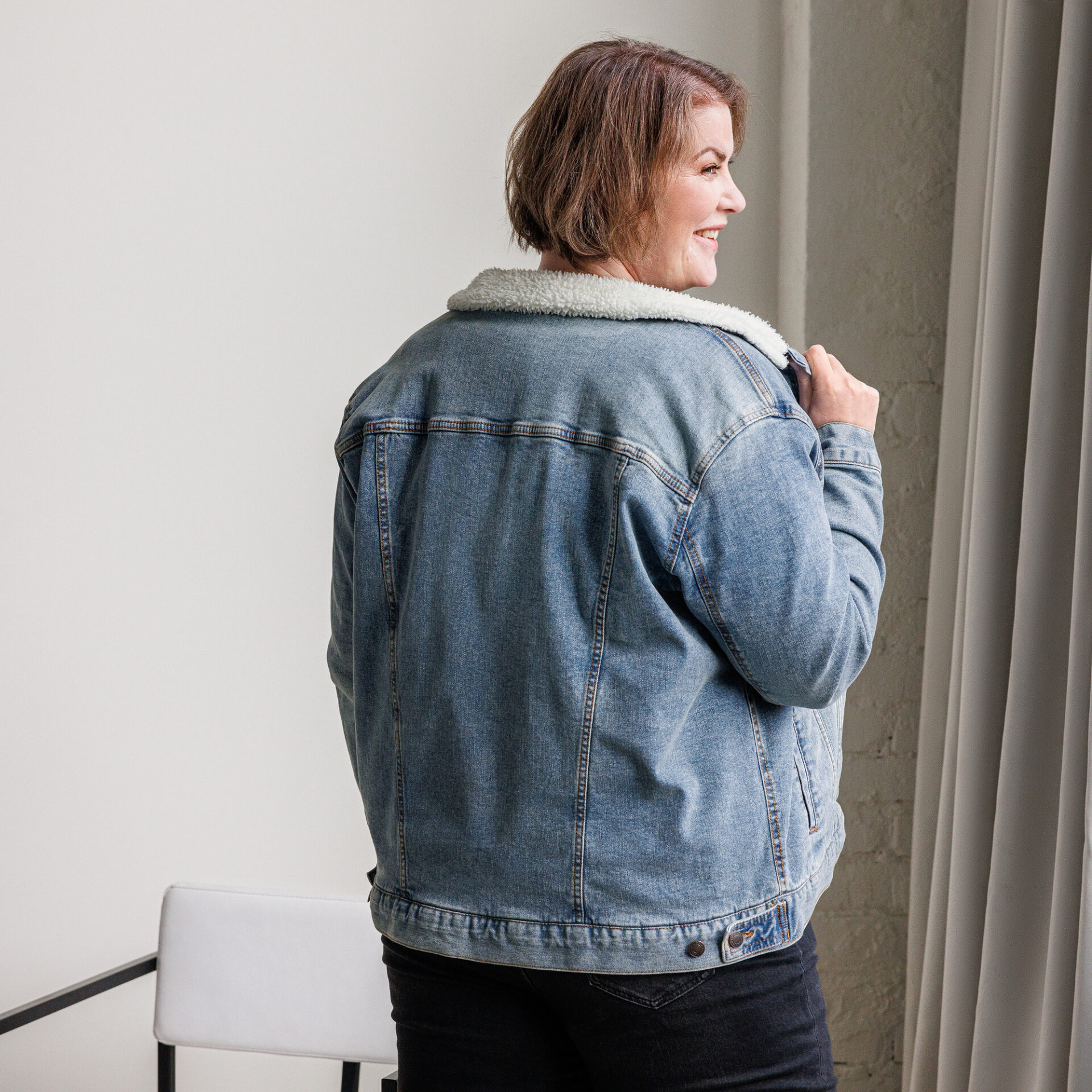 Woman in denim jacket standing by window.